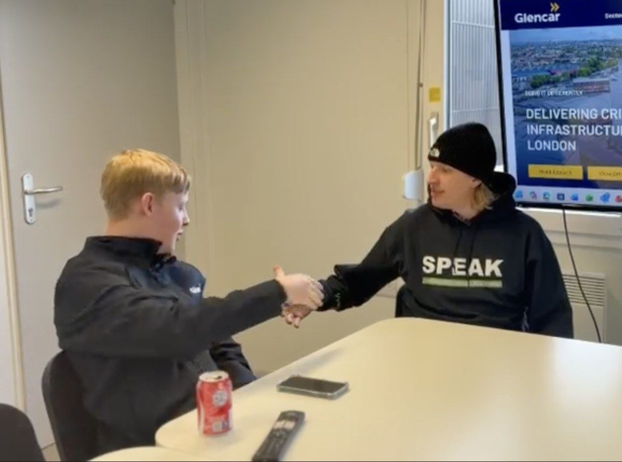 A SPEAK team member in a SPEAK-branded hoodie and beanie shaking hands with a young site worker across a meeting table, with a Glencar presentation screen behind them.