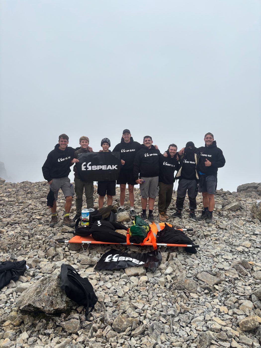 A team of SPEAK supporters in SPEAK hoodies holding a SPEAK banner at the summit of a misty mountain, part of a When One Falls community challenge climb.
