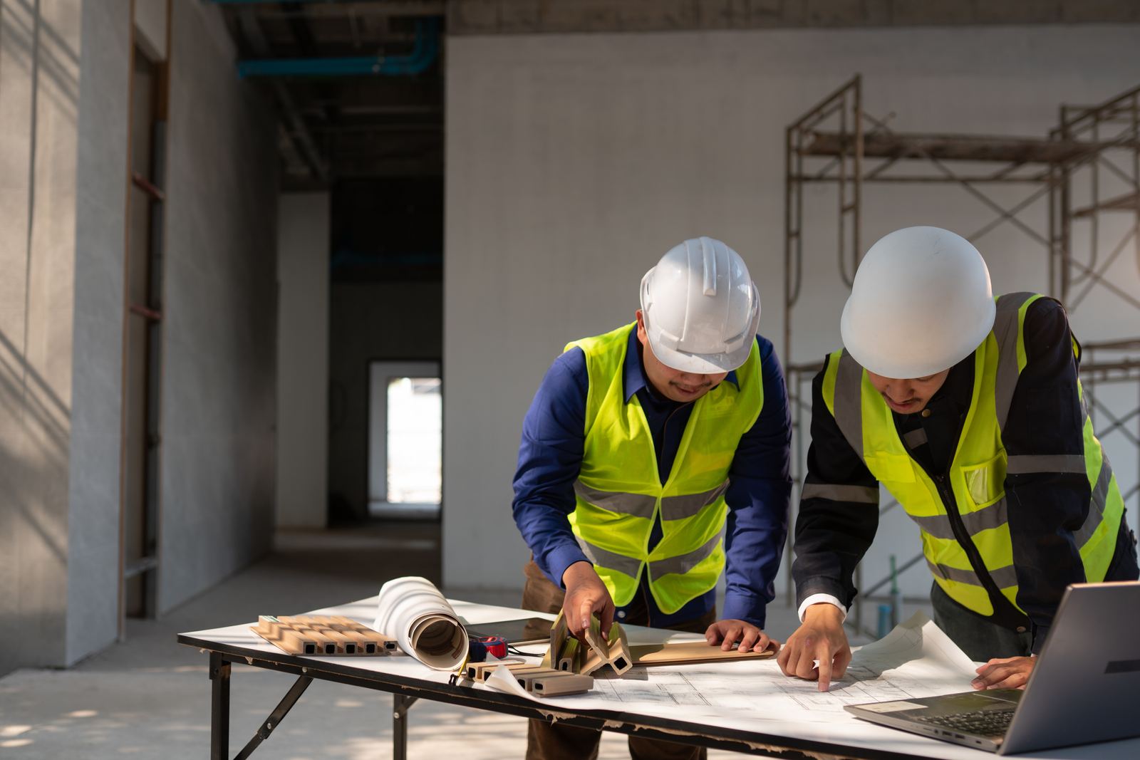 Two site leaders in hard hats and hi-vis vests reviewing building plans and a laptop together at a table inside an active construction site