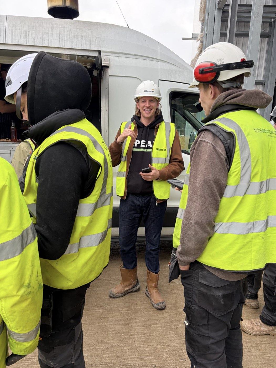 Alfie Ruffell, in a SPEAK hoodie and hi-vis, speaking with construction workers on site next to a Glencar van.