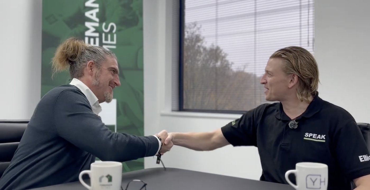A SPEAK team member in a SPEAK polo shirt shaking hands with a partner in an office meeting room.