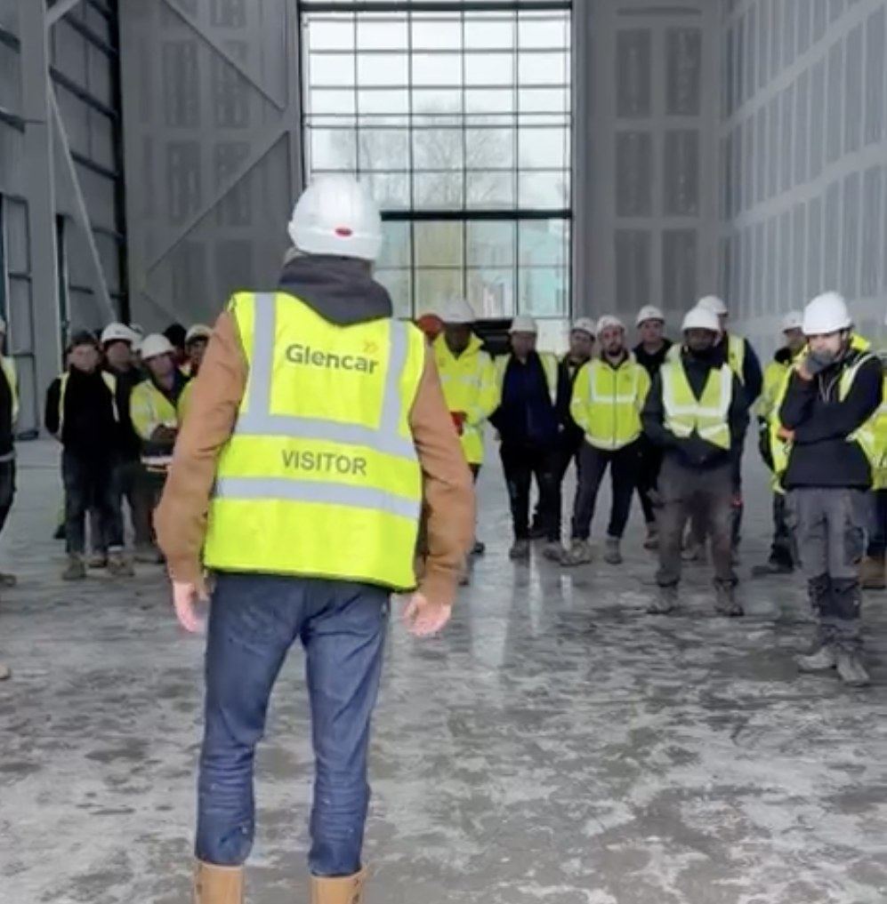 A Glencar site visitor in a hi-vis vest and hard hat, seen from behind, addressing a large group of construction workers gathered indoors on site.