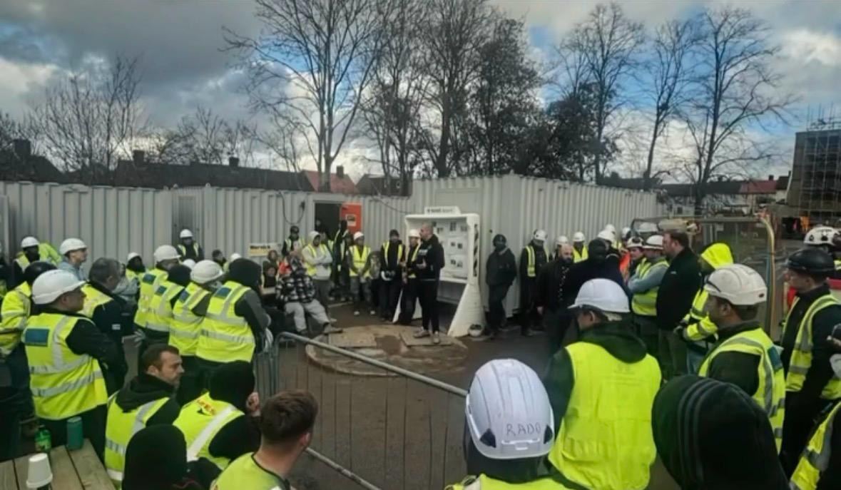 A large group of construction workers in hi-vis vests and hard hats gathered outdoors at a site cabin for a briefing.