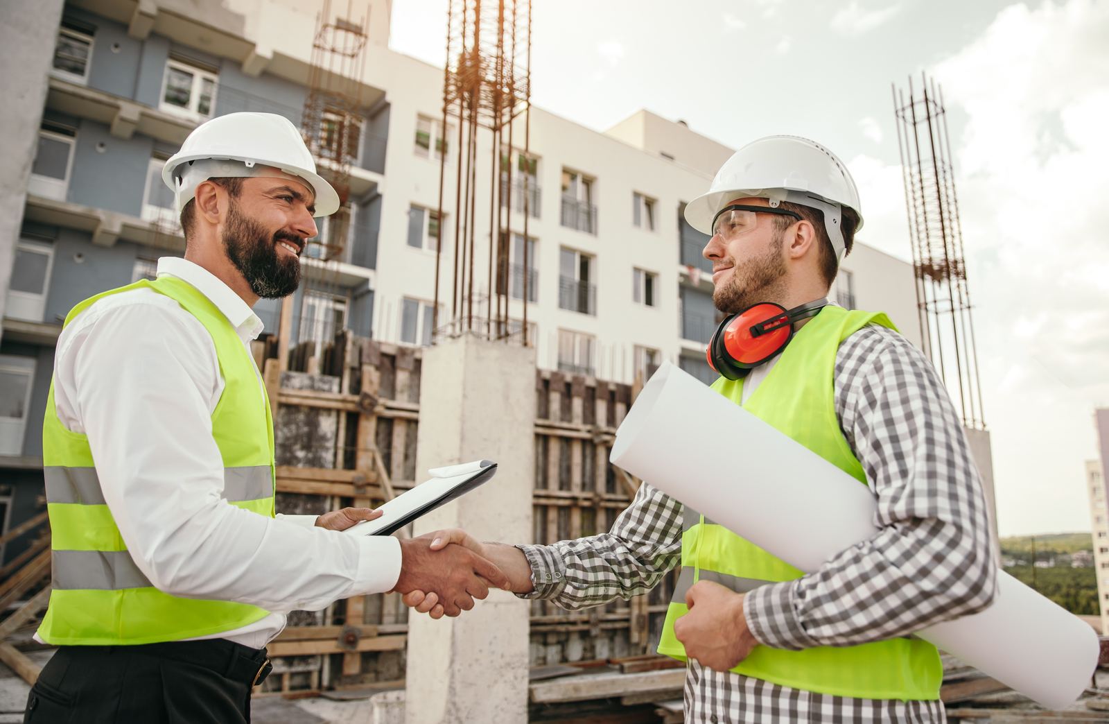 Two construction professionals in hi-vis vests and hard hats shaking hands on site, one holding a clipboard and the other holding rolled blueprints, with a building under construction behind them.