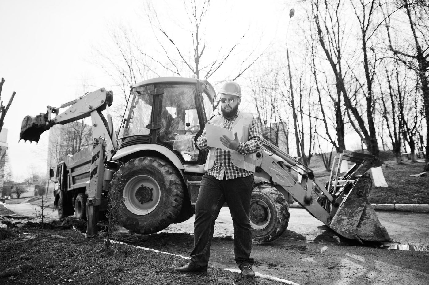 A construction worker in a hard hat and hi-vis, holding papers, standing in front of a digger on site - documentary-style construction storytelling.