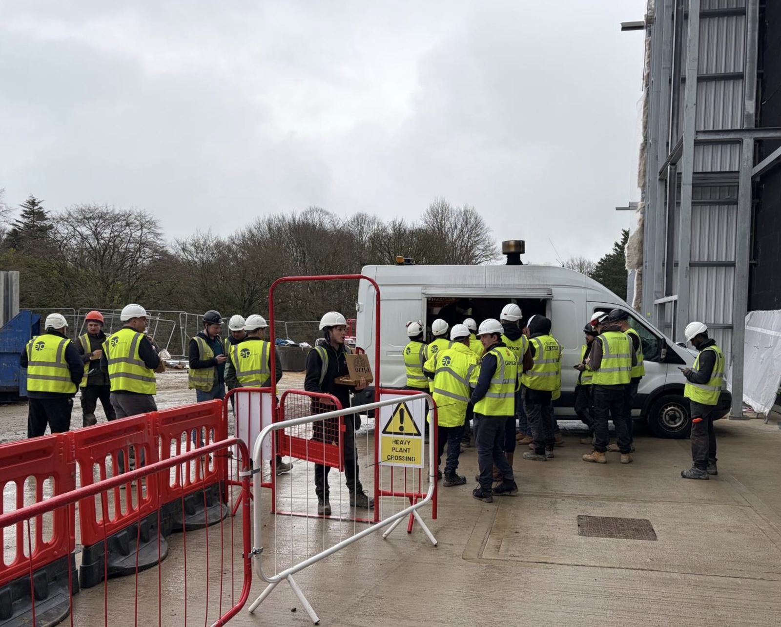 A group of construction workers in hi-vis vests and hard hats gathered around a site van during a break, with barriers and a heavy plant crossing sign in the foreground.
