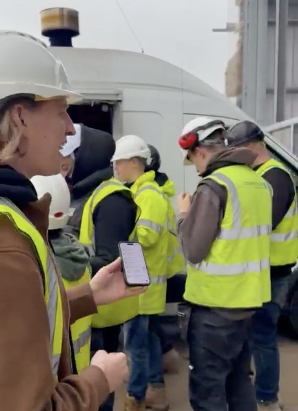 A SPEAK team member in a hi-vis vest and hard hat holding a phone, talking with a group of construction workers in hi-vis vests on site.