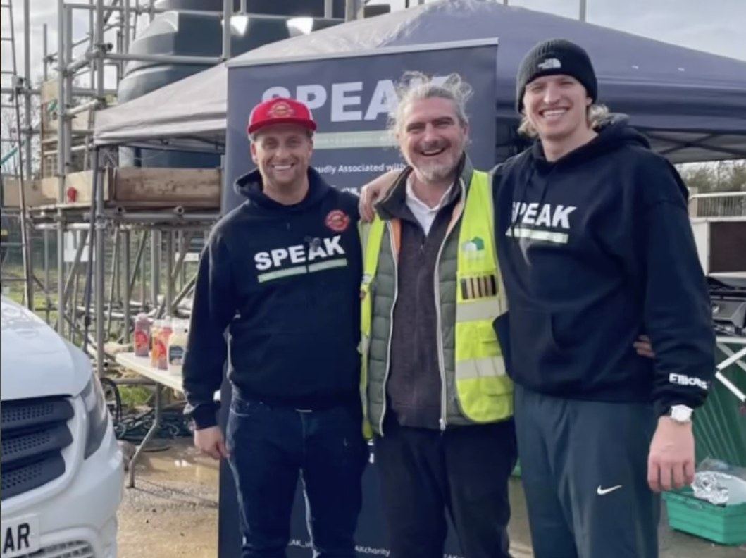 Three men smiling together at a SPEAK site event, two wearing SPEAK-branded hoodies and one wearing a hi-vis vest, in front of a SPEAK banner and scaffolding.
