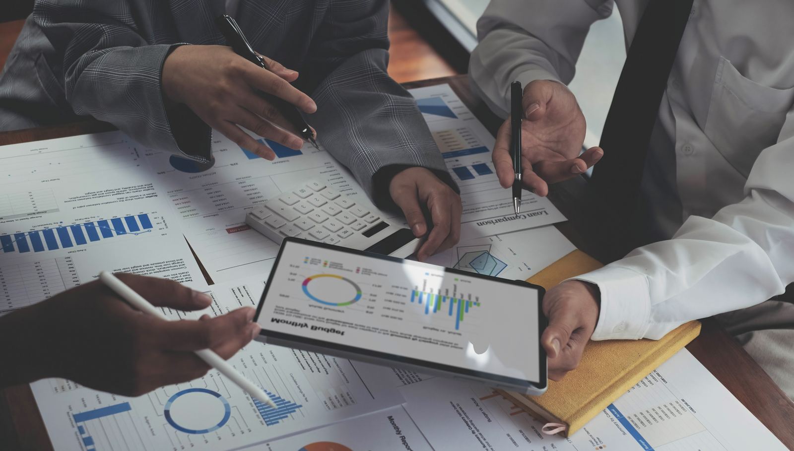 Colleagues at a table covered in printed charts and reports, reviewing a tablet showing pie and bar charts, illustrating a report and roadmap review.