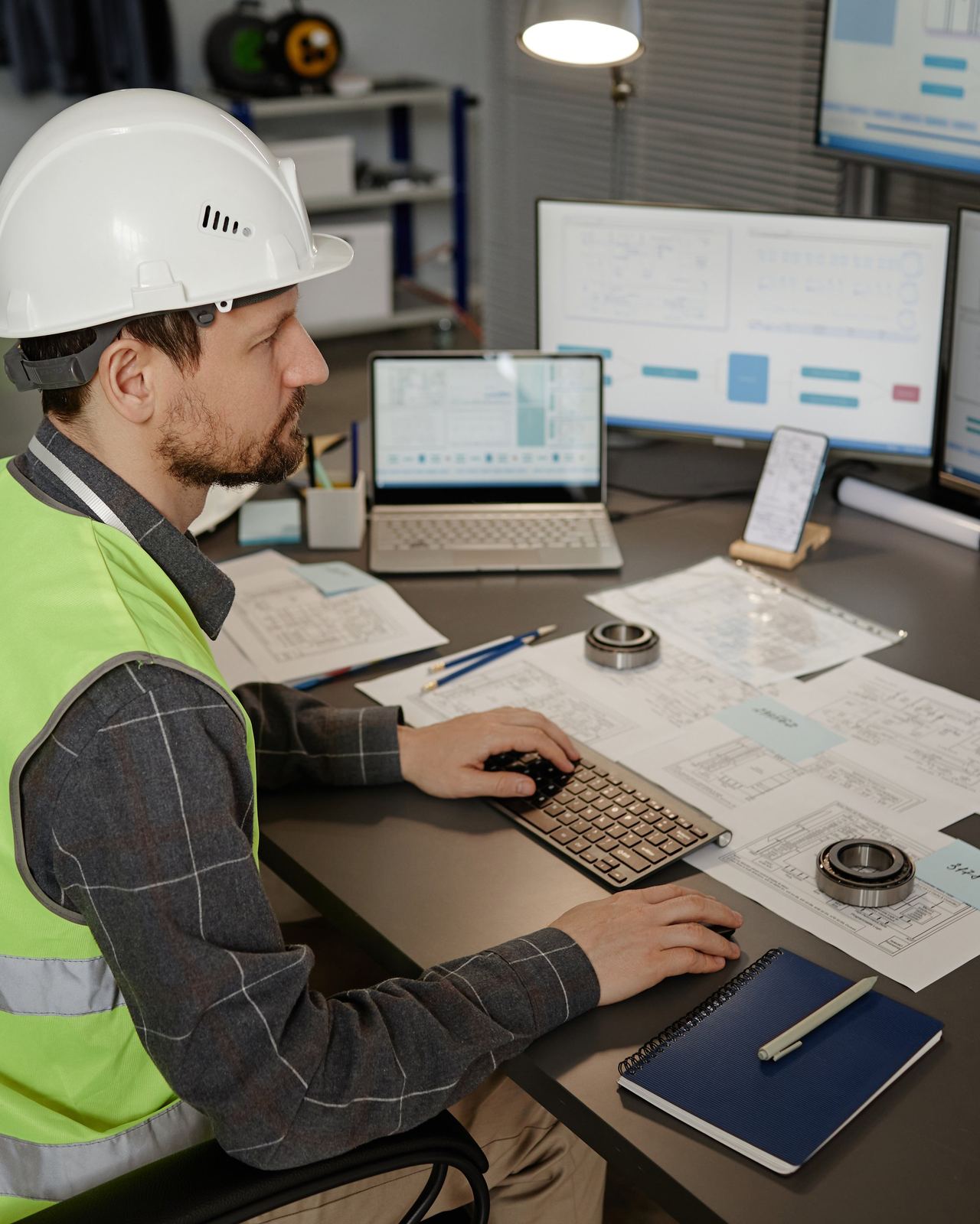 An engineer in a hard hat and hi-vis reviewing technical drawings and data across multiple screens at a desk