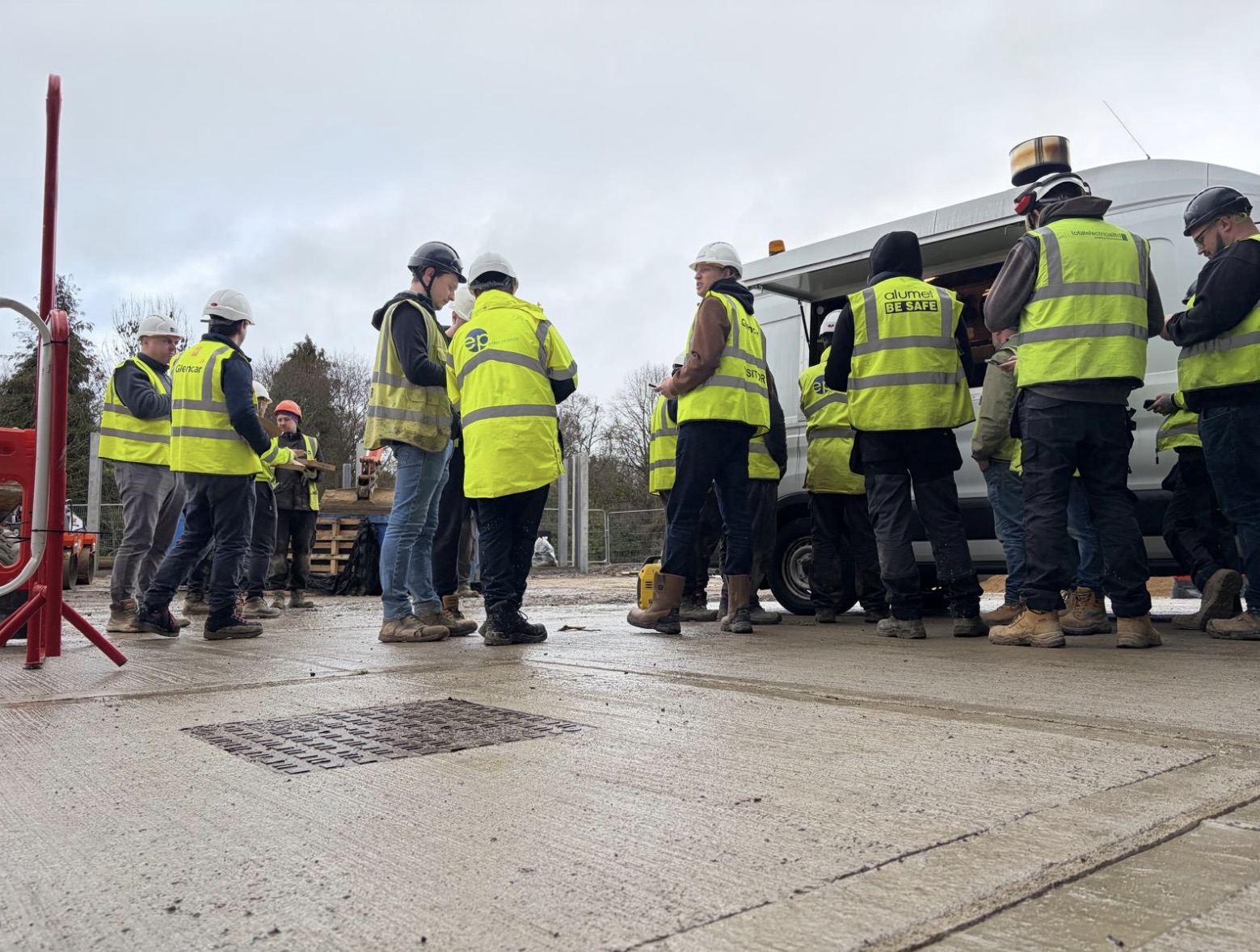 A group of construction workers in hi-vis and hard hats gathered round a site van talking during a toolbox talk